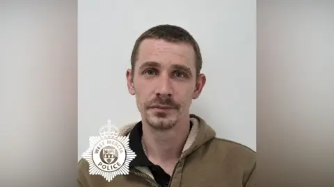 A police custody image of a man with short dark hair and short facial hair, wearing a green hoodie and stood in front of a light-coloured wall. There is a West Mercia Police logo in the bottom left corner.