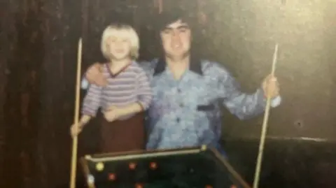 Hunter family A young boy and his dad pose for a photo with snooker cues in hand. A small snooker table is in front of them. The boy has long blond hair and is wearing a hooped shirt. His dad is wearing a blue shirt with a navy collar and has black hair.