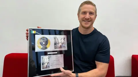 Lily-May Symonds/BBC Anthony Lanni, holding his platinum disc that shows artwork for (What's the Story) Morning Glory? 