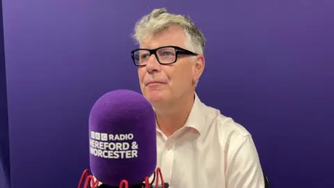 A man wearing glasses and a white shirt, with grey hair, is in front of a purple background. A BBC Hereford & Worcester radio microphone is in front of him.