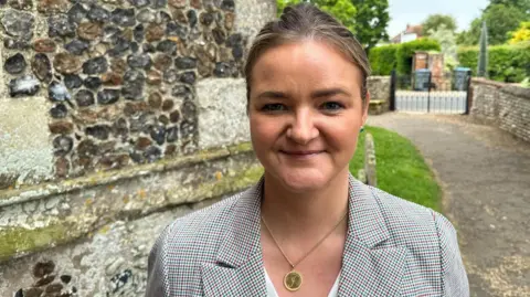 Ben Parker/BBC Jenny Riddell-Carpenter MP wearing a checked jacket and a white top stands in front of a church. She has brown hair, which is tied back, and is wearing a gold pendant. She is smiling at the camera.