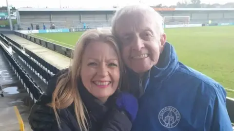 Colin Brookes smilining in a long blue coat. He is hugging a blonde woman who wears a longline black padded coat