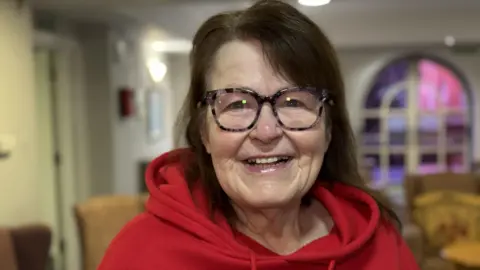 A woman with long dark hair, glasses and a red hooded top smiles as she stands inside a house.
