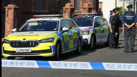 Police cars on a street in front of a house with police tape in the foreground and uniformed officers in background