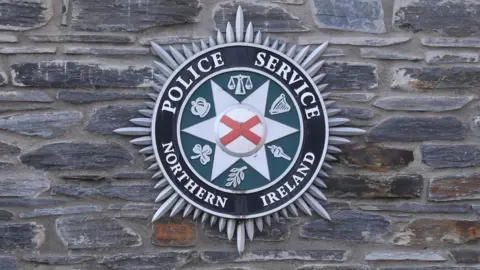 The PSNI badge on a stone wall. The badge is a Bath star with lettering around the inside reading "police service of Northern Ireland". In the centre is a red and white St Patrick's Saltire.