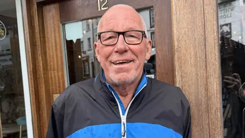 Colin Ball wearing glasses and a black-and-blue zip-up jacket stands facing the camera in front of a wooden doorway. The door has the number 12 above it, and parts of nearby buildings are visible through the glass panels on either side