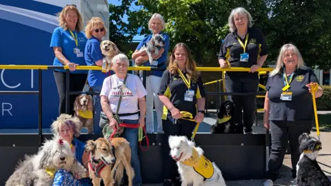Eight women with lanyards wearing blue, black or white T-shirts. They are all holding dog leads. The dogs include an old English sheepdog, a Saluki, two spaniels and a Tibetan terrier.