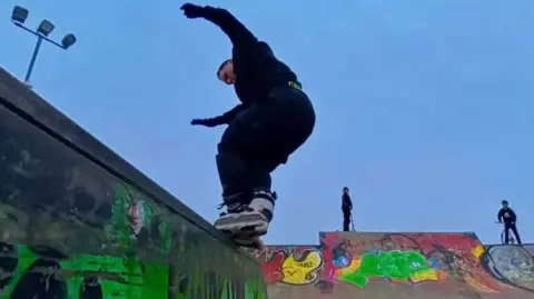 A man on roller blades, wearing a black shirt and black trousers performing a stunt on a wall. Two people can be seen in the background on scooters. The lights on top can be seen switched off while the place looks dark.