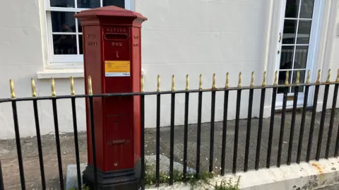 Red hexagonal Victorian post box next to black and gold railings and in front of a cream coloured building with a white sash window and door visible.