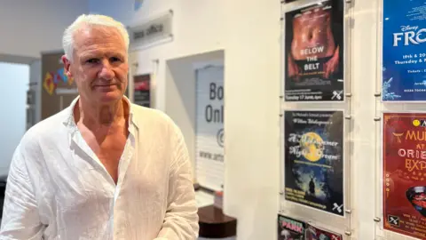 Tom Binet has gray hair, wearing a white shirt, stands in front of a wall displaying various event posters. The posters include titles like 'Below the Belt,' 'Frozen,' and 'Music at the Orangerie.' Above the posters is a sign that reads 'this week.' In the background, there is a box office sign and another poster partially visible through an open doorway.