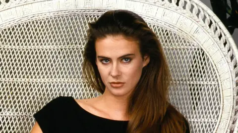 Annabel Schofield, a woman with long brown hair sits with in a white wicker chair, wearing a black dress. She looks directly at the camera with a serious expression.