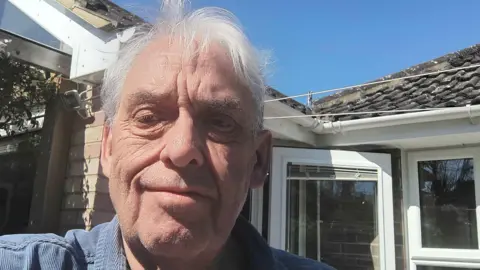 Graham Turner Graham Turner with short white hair smiling at the camera and wearing a blue T-shirt. He is standing in front of a white glazed door and white-framed windows on a brick house. There is a blue sky above.