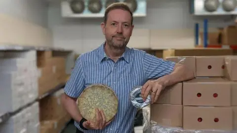 Simon Spurrell Simon Spurrell is posing with a wheel of blue cheese next to some boxes. He is wearing a blue and white striped linen short sleeved shirt.