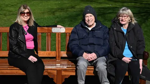 Greg Macvean Three people sit on a bench. On the right is a lady with blonde hair and wearing sunglasses next to her is a man wearing a large navy jacket and a hat as well as another woman wearing a black jacket and glasses.