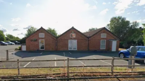 BBC A fence and grass are in the foreground and a building is in the background. Trees are behind it on the photo. The building is made of three connected and arched buildings next to each other, made of red brick with white-framed windows.
