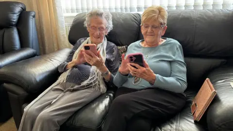 Simon Thake/BBC Two elderly women sit together on a sofa smiling as they look down at their mobile phones