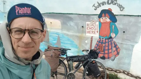 Christoph Held is wearing a navy "Plantur" cap and a light blue coat. His bike is behind him leaning against a wall with a painting of a ginger haired man wearing a kilt holding a sign saying "Road ends here". Above the painting are the words "John O'Groats".