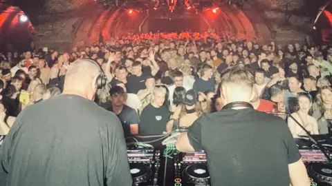 Soma Records Two men in black t-shirts DJ on CDJs in front of a full audience in a nightclub