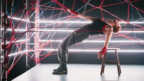 Kirsten McTernan A performer dressed in black on stage in the centre of a web of red, blue and white rope