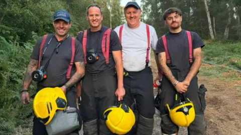 Four firefighters stood in a forest smiling at the camera. They have their protective black trousers on with red ties that go over their shoulders, and they are carrying yellow helmets. Three of them have black T-shirts on, and the other is wearing a white one. Two of them are wearing caps.