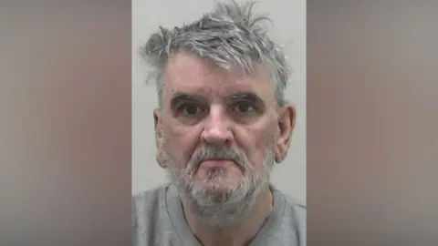 Northumbria Police Mugshot of David Elliot. He has thick scruffy grey hair and an untidy grey beard