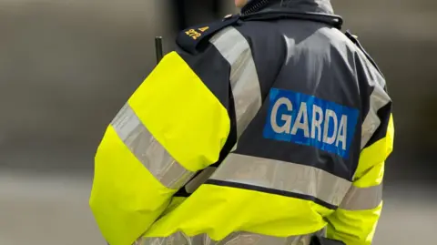 The back of a gardaí uniform raincoat. Garda is wrriten in white and blue, and the rest of the coat is a reflective neon yellow colour with silver stripes on the arms and chest area.
