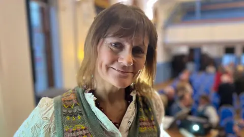 A woman with light brown hair, wearing a white blouse and green tank top, looks at the camera