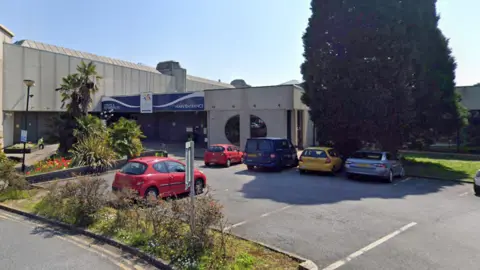 Google A photo of the outside of the vaccination centre. It is a big building with a blue entrance sign above the door. There is a car park outside with lots of cars in it. There is also plenty of greenery and a big tree outside. 