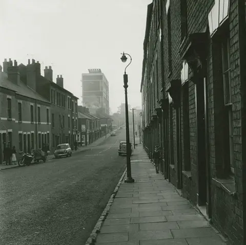 Estate of John Blakemore provided by Coventry University A street scene of a 1960s terraced road in Coventry with a few cars parked on either side of the road and black lamp posts sporadically placed along the pavement