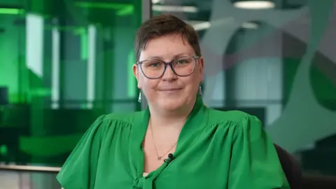 Aimee is wearing a green blouse and is smiling at the camera. She is wearing glasses and has short, dark hair. She is in an office setting, with green clear panels behind her.
