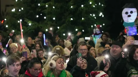 City of Wolverhampton Council Crowds gather in the street in Bilston. Christmas trees can be seen at the back of the images with lights on then a large crowd of people, some dressed festively, many holding little lights or have their phones held in the air. A decorative snowman can be seen on the right of the image.