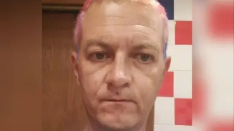 US Secret Service Aleksej Besciokov with pink and purple coloured hair standing in front of a white wall with red and blue tiles.