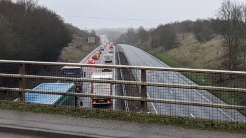 BBC A view of the A47 from a bridge running above it. The left hand carriageway is filled with cars travelling away from the camera in long queues. The right hand carriageway is empty due to the closure.