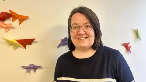 BBC News A lady with glasses and short hair smiling next to a wall of origami butterflies