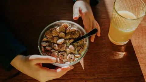 Top down image of a little boy sitting at a sunny dining table and eating a small bowl of breakfast cereal. A glass of fresh orange juice sits off to one side.