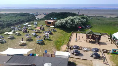 Discover Brean A view from above of the outdoor seating area at the Sundowner Beach Bar, overlooking Berrow beach. There are lots of wooden benches with parasols set up on the grass, and a children's sandy play area on the right with a wooden playhouse and swing set. The beach and the sea are visible in the distance, past the grassy dunes.