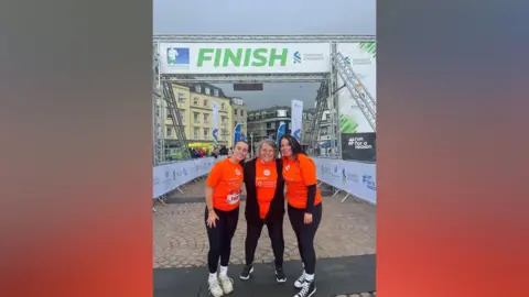 Josie Carey All three smile at the camera at the finish line of the Jersey Marathon. They're wearing bright orange tops and black running leggings.