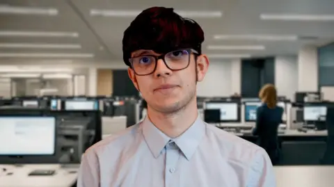 Ben Moore/BBC Tommy Hills is wearing dark framed glasses and a pale shirt. Behind him are rows of computer screens