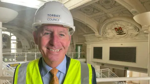 Chris Lewis is inside an upper floor of the Pavilion, which has an ornate roof. He smiles and is wearing a hard hat, with stairs behind him.