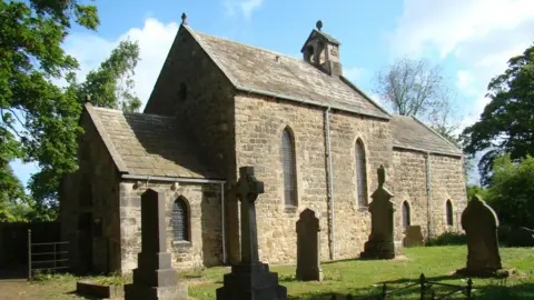 Church of Our Lady, from the South West. It is a small stone church with graves in the grassy yard.