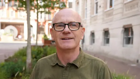 Qays Najm/BBC A man wearing black frame glasses and a dark green shirt with his top button undone. He is looking at the camera and has a faint smile. He is standing outside 