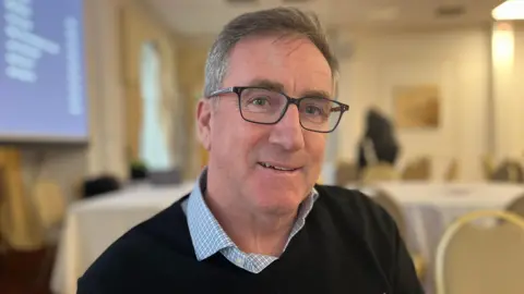 Olivia Fraser/BBC Dressed in a black jumper and shirt with a pale blue and white check and glasses Nick Brown smiles at the camera.