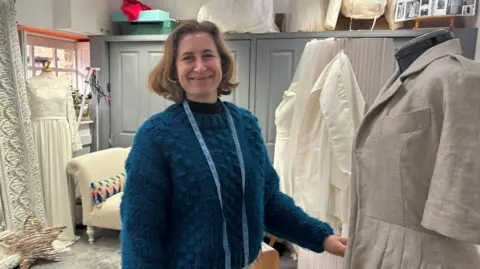 A mid-shot of Amy in a fluffy blue jumper with a tape measure round her neck. She is in her shop with a mannequin in the foreground wearing a linen dress. There are other mannequins in the background with wedding dresses on and other props and an album of photographs on a high shelf
