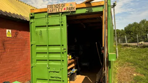 A green shipping container with the door open and a sign reading "friends of bartley park" above it.
