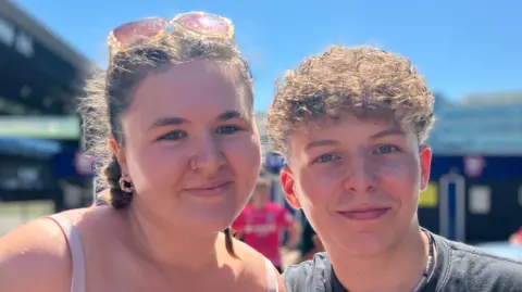 Alice Cunningham/BBC Caitlin Brundle and Thomas Portway smile at the camera. Caitlin has brown hair that has been braded and Thomas has short brown curly hair.