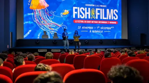 Two people talk to a room full of cinema goers sat in red chairs, with a blue screen and Fish and Films logo projected onto the screen behind.