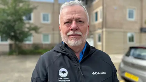 Steven Tatlock outside the coroner's court. He has a neat short white beard and short white hair and is wearing a black jacket with the Lake District national Park logo on the breast.