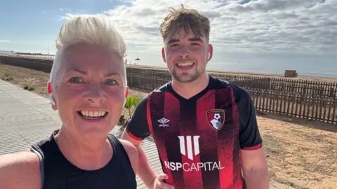 Children with Cancer Two people pose for a selfie on a seaside path, one wearing a sleeveless running top and the other a red and black football shirt, with sand, fencing and the sea visible behind them under a cloudy sky.