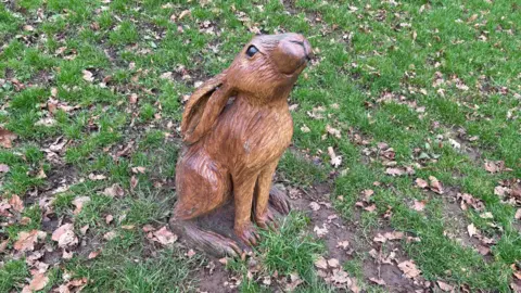A carved wooden hare sitting on a patch of grass in winter. The statue has long ears tucked back, large brown eyes and it is looking up towards the sky.