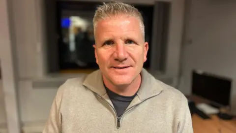 Chris Quinn looking at the camera he has grey hair and is wearing a grey quarter zip and navy top.